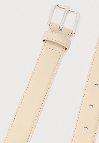 Cinturón de cuero beige con una textura suave, hebilla plateada y agujeros de ajuste espaciados uniformemente a lo largo de la correa. Detalles de costuras visibles.