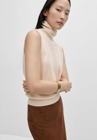 Gebreide beige mouwloze coltrui met een aansluitende zoom, gecombineerd met een bruine suède rok. Een gouden armband wordt om de pols gedragen.