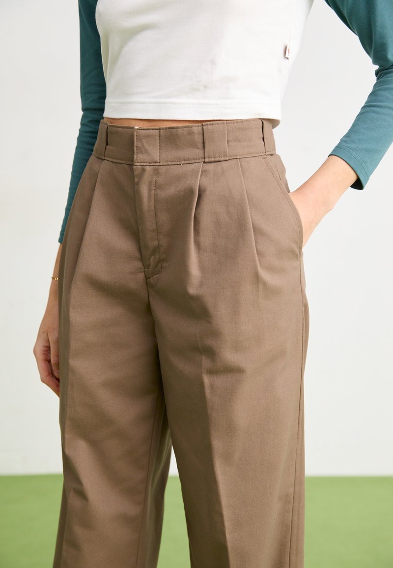 Braune, taillierte Hose mit hoher Taille, plissiertem Vorderteil, Seitentaschen und glattem Stoff. Kombiniert mit einem weißen Crop-Top und türkisfarbenen Ärmeln.