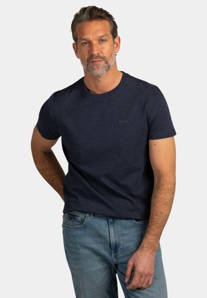 Man met kort grijs haar en baard, gekleed in een marineblauw T-shirt en lichtblauwe spijkerbroek, staand met één hand achter zijn rug.