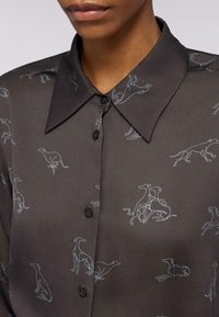 Nahaufnahme eines schwarzen Hemdes mit spitzem Kragen und hellblauen Zeichnungen von sitzenden und stehenden Windhunden.