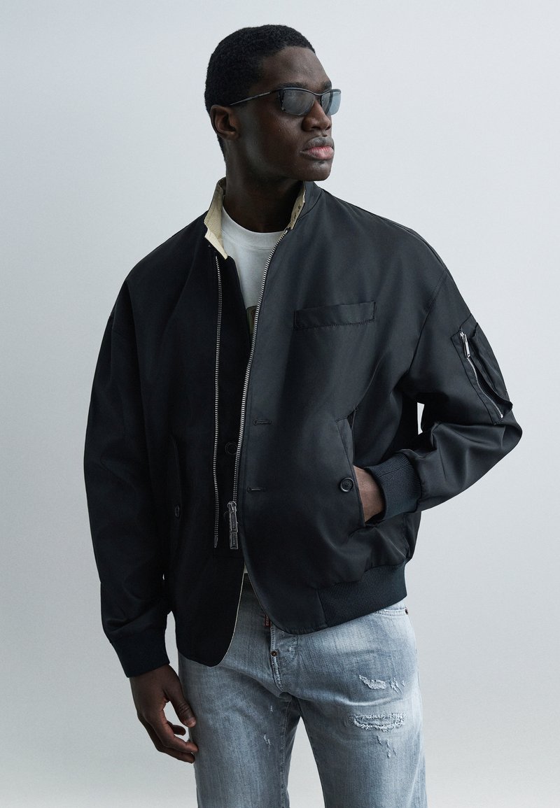 Homme portant une veste bomber noire, une chemise blanche, un jean bleu clair déchiré et des lunettes de soleil foncées, regardant sur le côté avec une main dans la poche.