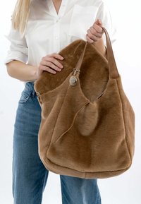 Sac en peluche marron avec une texture douce, des accents en cuir et une étiquette en argent. Grande forme décontractée avec une large ouverture et une seule poignée.