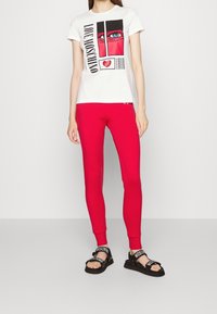 Biała koszulka z grafikom przedstawiającym oko i serce, zestawiona z dopasowanymi czerwonymi legginsami i czarnymi sandałami z logo na paskach.