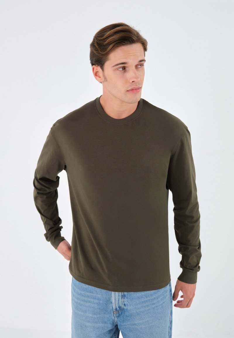 Langärmliges Rundhals-Shirt aus dunkelolivgrüner Baumwolle. Hat einen lockeren Schnitt mit hochgerollten Ärmelabschlüssen. Kombiniert mit hellblauen Denim-Jeans.