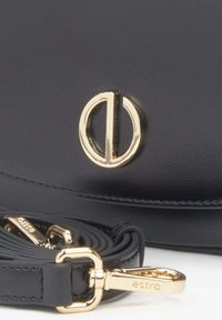 Černá kožená taška s hladkou texturou, s kulatým zlatým logem, odnímatelným popruhem a leštěnými zlatými detaily.