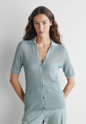 Femme aux longs cheveux bruns portant un cardigan boutonné à manches courtes bleu clair et un pantalon assorti, debout contre un mur blanc.