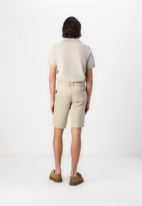 Beige kurzärmliges, strukturiertes Hemd mit Streifen, kombiniert mit hellbeigen Shorts. Braune, flache Schuhe runden das Outfit ab.