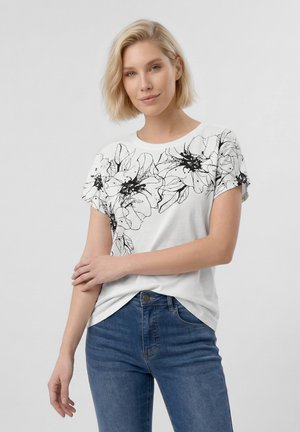 Szőke nő fehér pólóban, amelyen nagy fekete virágminta díszíti a mellrészt, és kék farmert visel, karját keresztbe téve áll.