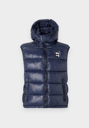 Marineblå puffervest med hette, glossy materiale, glidelås, to frontlommer og et logo-patch på venstre bryst.