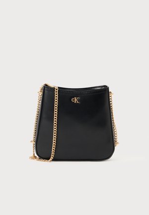 Sac à main en cuir noir avec bandoulière chaîne dorée et petit logo doré "cK" sur le devant, sur un fond clair uni.
