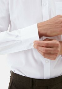 Witte lange mouwen shirt met knoopsluitingen, gladde stof en subtiele glans. Handen die de manchet aanpassen bovenop geblokte bruine broek.