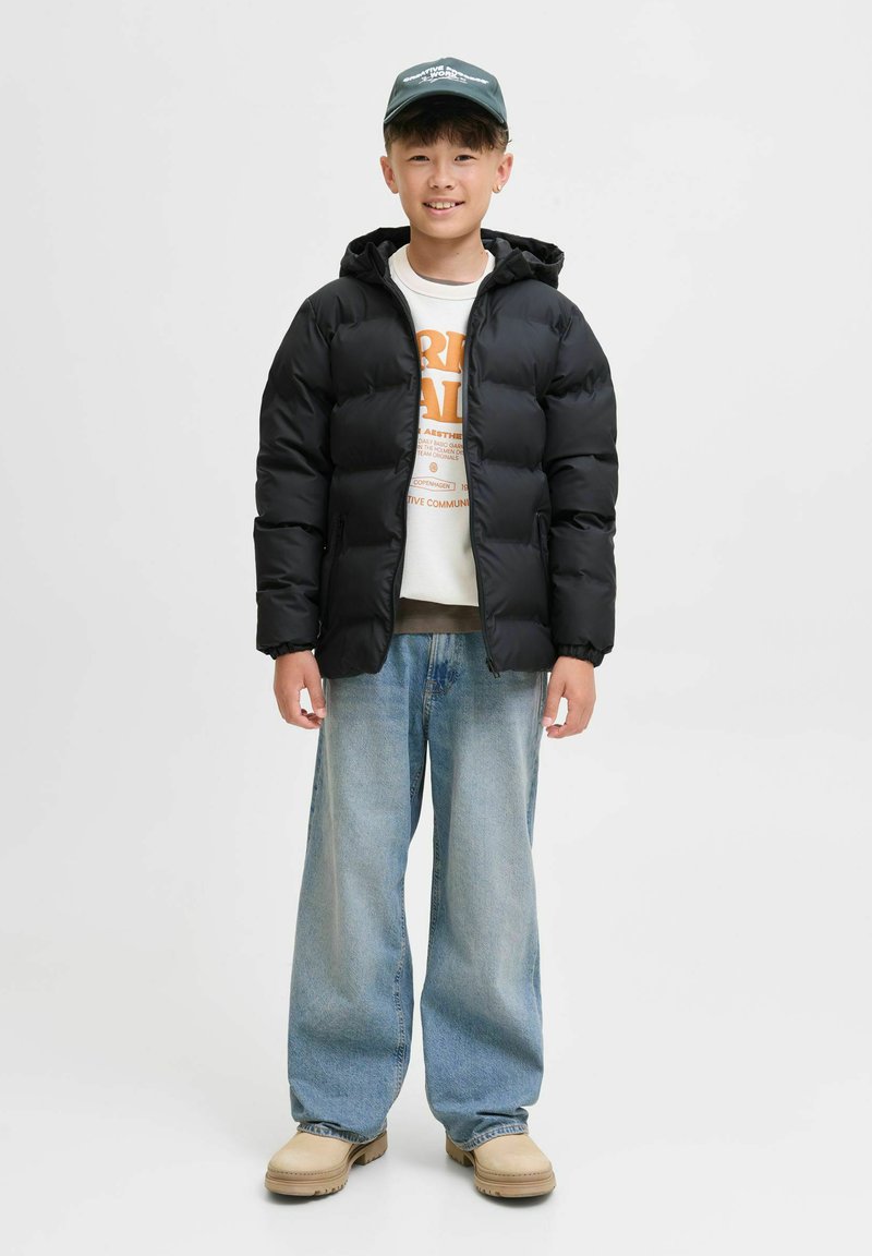 Doudoune noire avec capuche, fermeture éclaire à l'avant et poignets élastiques sur un pull blanc. Jean léger bleu clair à coupe ample et chaussures beiges.