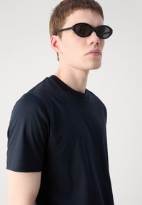 T-shirt nera a maniche corte con scollo rotondo. Modello che indossa occhiali da sole neri a forma angolare, su uno sfondo grigio chiaro.