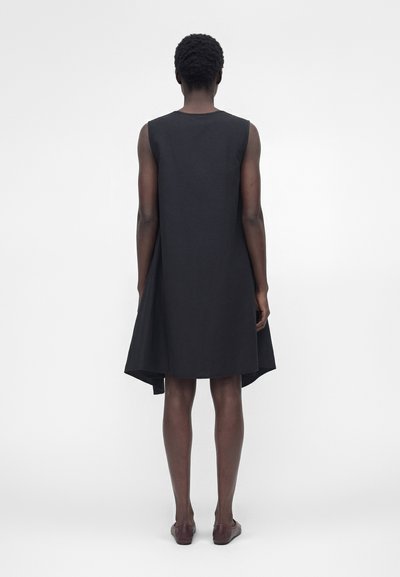 Γυναίκα με κοντά σγουρά μαλλιά, που φοράει ένα μαύρο ασύμμετρο φόρεμα χωρίς μανίκια και καφέ παπούτσια, στέκεται γυρισμένη με την πλάτη σε λευκό φόντο.