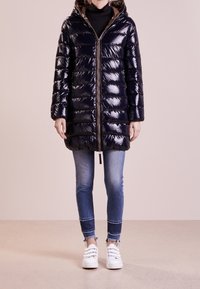 Lyst marinblå puffjacka med huva, med dragkedja fram och horisontell quiltning, kombinerad med blå jeans och vita sneakers.