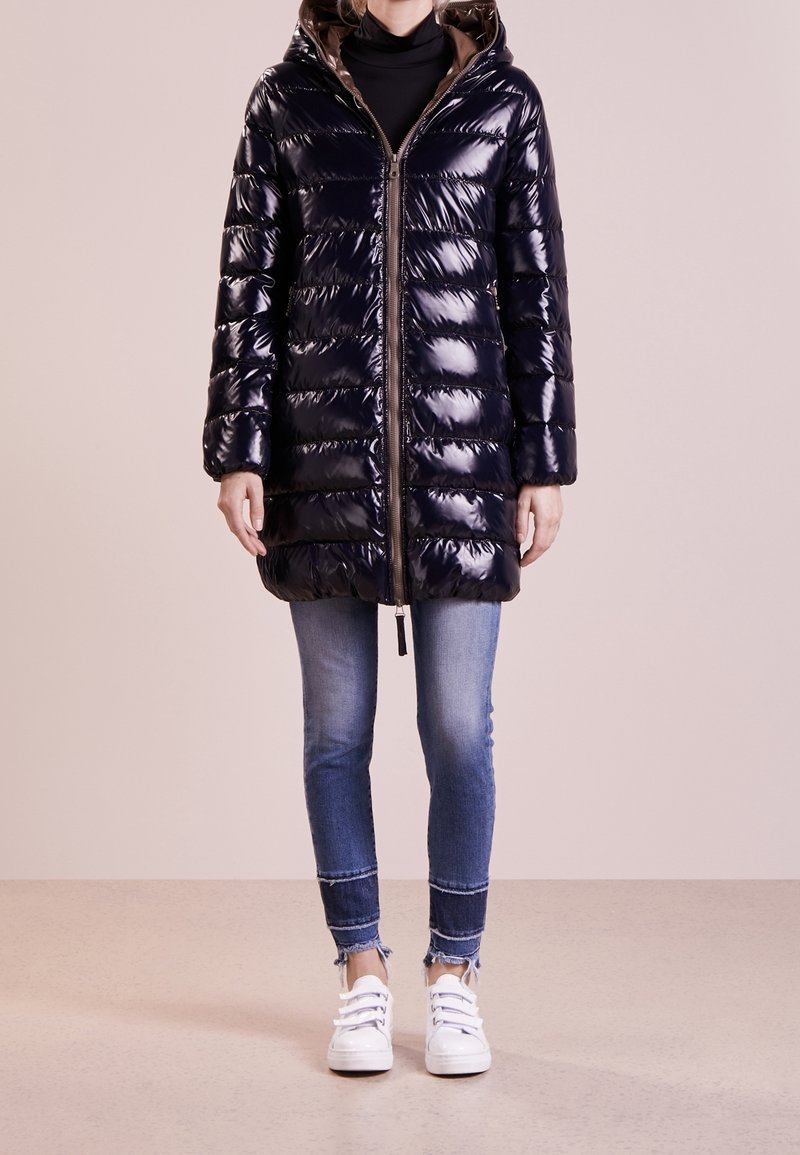 Lyst marinblå puffjacka med huva, med dragkedja fram och horisontell quiltning, kombinerad med blå jeans och vita sneakers.