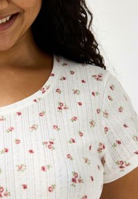 Bílý top s krátkými rukávy s texturovaným žebrovaným vzorem a rozptýlenou růžovou květinovou výšivkou. Kulatý výstřih s jemnými zdobeními na tkanině.