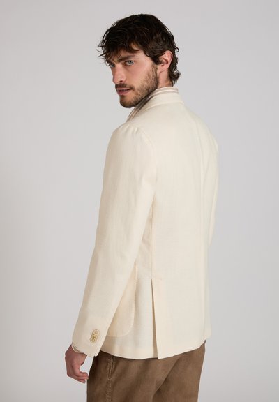 Man met kort krullend haar en baard, gekleed in een crèmekleurig colbert en een bruine broek, kijkt over zijn schouder tegen een effen achtergrond.