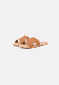 Brown slide sandals with two wide straps featuring cut-out designs. The sole is flat and made of a light, natural material.