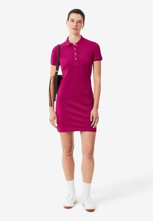 Frau trägt ein kurzärmeliges magentafarbenes Polo-Kleid, weiße Socken, weiße Turnschuhe und trägt eine braune Umhängetasche, steht vor einem einfarbigen Hintergrund.
