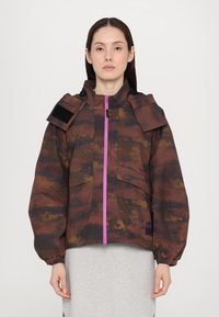 Braun camouflierter Jacke mit Kapuze, oversized Schnitt und elastischen Manschetten. Verfügt über einen lila Reißverschluss und Schulterlaschen für zusätzliche Details.