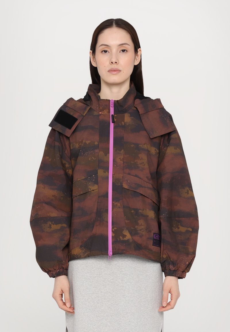 Braun camouflierter Jacke mit Kapuze, oversized Schnitt und elastischen Manschetten. Verfügt über einen lila Reißverschluss und Schulterlaschen für zusätzliche Details.