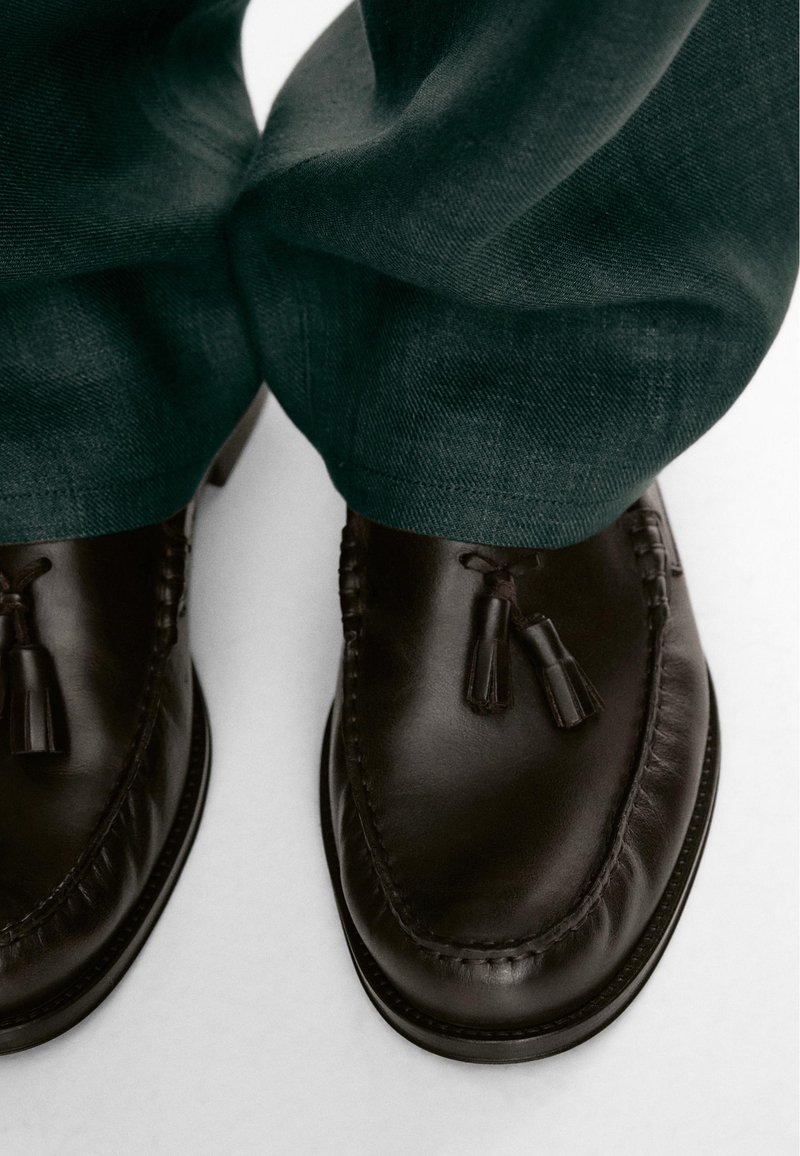 Mocassini con nappine in pelle marrone scuro indossati con pantaloni verde scuro, visti dall'alto che mostrano dettagli delle scarpe e la trama del tessuto.