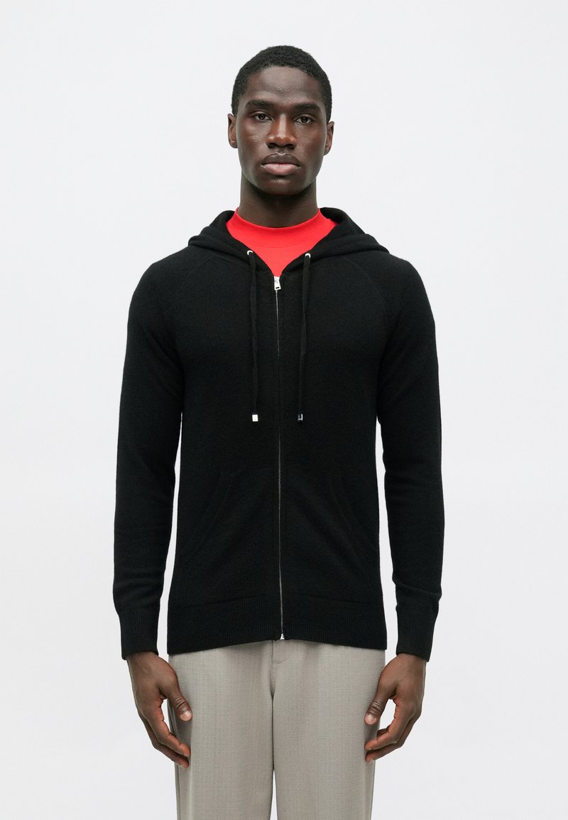 Homem a usar um hoodie preto com fecho, uma camisa vermelha por baixo e calças cinzentas claras, de pé contra um fundo branco liso.