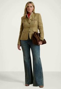 Bézs színű, strukturált blézer, két mellzsebbel, kék, szárán szűkülő farmernadrággal párosítva. A modell egy barna, láncos pántú kézitáskát tart.