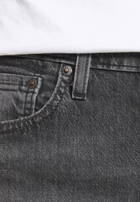 Acercamiento de un bolsillo de jeans de mezclilla gris oscuro con remaches metálicos y costuras, combinado con una camisa blanca Metropolitana.