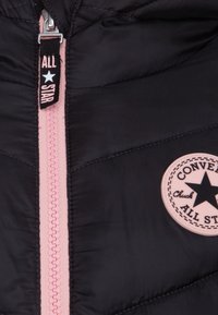 Doudoune noire avec une fermeture éclair rose, un patch rond rose portant l'inscription "Converse Chuck All Star" et une texture matelassée.