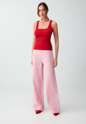 Femme debout portant un haut sans manches rouge, un pantalon large rose clair, des chaussures rouges et une écharpe à motifs autour du cou sur un fond uni.