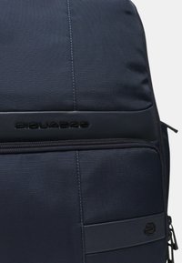 Zaino blu navy con tessuto testurizzato, design elegante, cerniera centrale e logo in rilievo. Include una tasca laterale e dettagli in metallo resistente.
