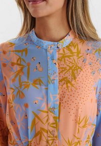 Blouse à manches longues avec boutons, de couleur bleu clair et pêche, ornée de motifs botaniques verts et jaunes, avec un col rond et une fermeture à boutons sur le devant.