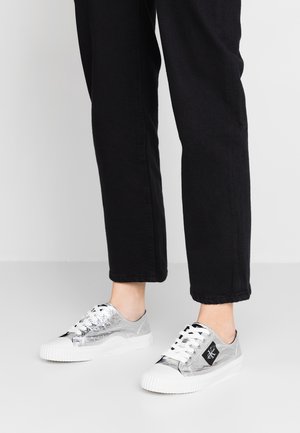 Persona que lleva pantalones negros y zapatillas plateadas metálicas con cordones y suelas blancas, de pie sobre una superficie gris.