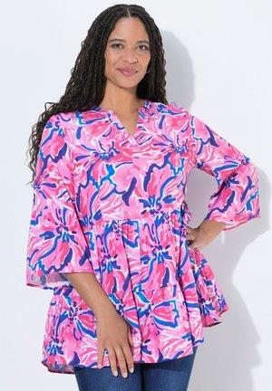 Frau mit langem geflochtenem Haar, die eine locker sitzende Bluse mit pinkem, blauem und lila Blumenmuster und blaue Jeans trägt, steht mit einer Hand in der Hüfte.