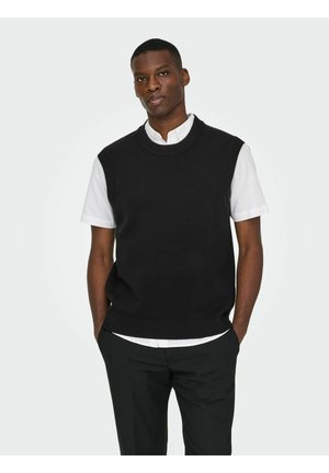 Gilet en tricot noir avec un col rond, porté sur une chemise blanche à col. Le modèle pose les mains dans les poches, présentant un design épuré et simple.