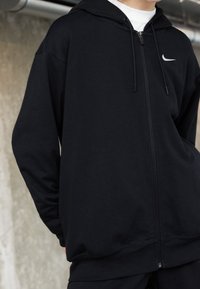 Svart zip hoodie i slätt tyg, med dragsko i huvan, sidofickor och en liten vit logotyp på vänster bröst.