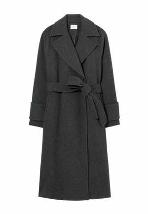 Donna in piedi indossa un lungo cappotto grigio scuro, camicia grigia, pantaloni larghi grigi, tiene una piccola borsa nera su uno sfondo grigio chiaro semplice.