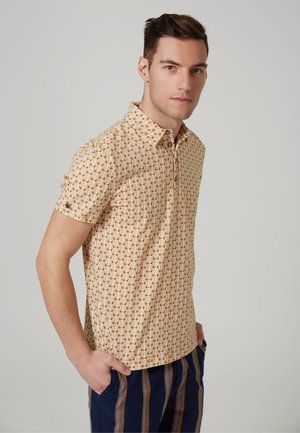 Camisa de manga corta en color beige claro con un patrón geométrico. Presenta un cuello abotonado y una textura suave. Combinada con pantalones a rayas.