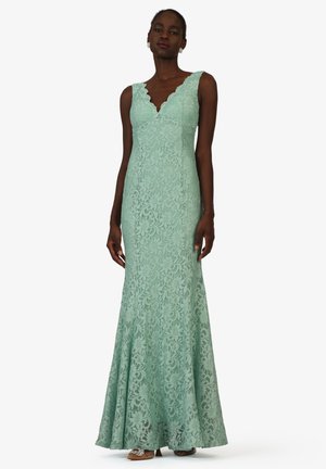 Woman wearing mint green sleeveless lace gown with scalloped V-neckline and flared hem, standing against plain white background.