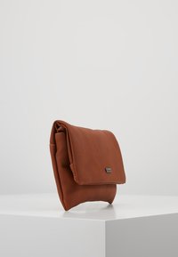 Borsetta a tracolla in pelle marrone con finitura texturizzata, pattina pieghevole e piccolo logo metallico. Posizionata su una superficie di esposizione bianca.