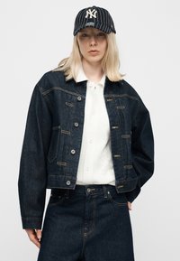 Ξανθός/ή φοράει μαύρο καπέλο New York Yankees με λεπτές ρίγες, σκούρο τζιν μπουφάν, λευκό πουκάμισο με κουμπιά και σκούρο τζιν παντελόνι, μπροστά σε απλό φόντο.