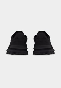 Paire de baskets de sport noires vues de dos, présentant des semelles texturées et des colliers de cheville rembourrés sur un fond blanc uni.