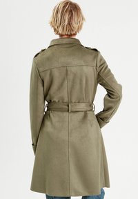 Abrigo trench de color verde oliva con cintura ceñida por un cinturón, mangas largas, hombreras y un acabado suave y texturizado. Vista desde atrás.