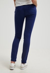 Marinblå slim-fit-byxor med en slät textur och diskreta bakfickor, kombinerat med vita slip-on-skor.