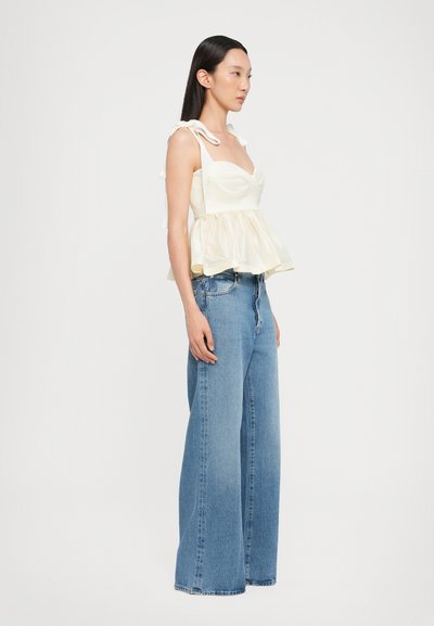 Top peplum blanco con tirantes atados, con un cuerpo texturizado, combinado con jeans de tiro alto y pierna ancha en azul. Tela suave, con un ligero brillo en la parte superior.
