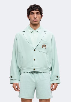 Mand iført lys mintgrøn knapjakke med broderet logo på brystlomme og matchende shorts, stående mod ensfarvet baggrund.