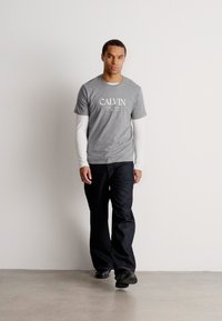 Mężczyzna idący w pomieszczeniu, ubrany w szary t-shirt Calvin na białe długie rękawy, czarne spodnie i czarne buty, na tle gładkiej białej ściany.